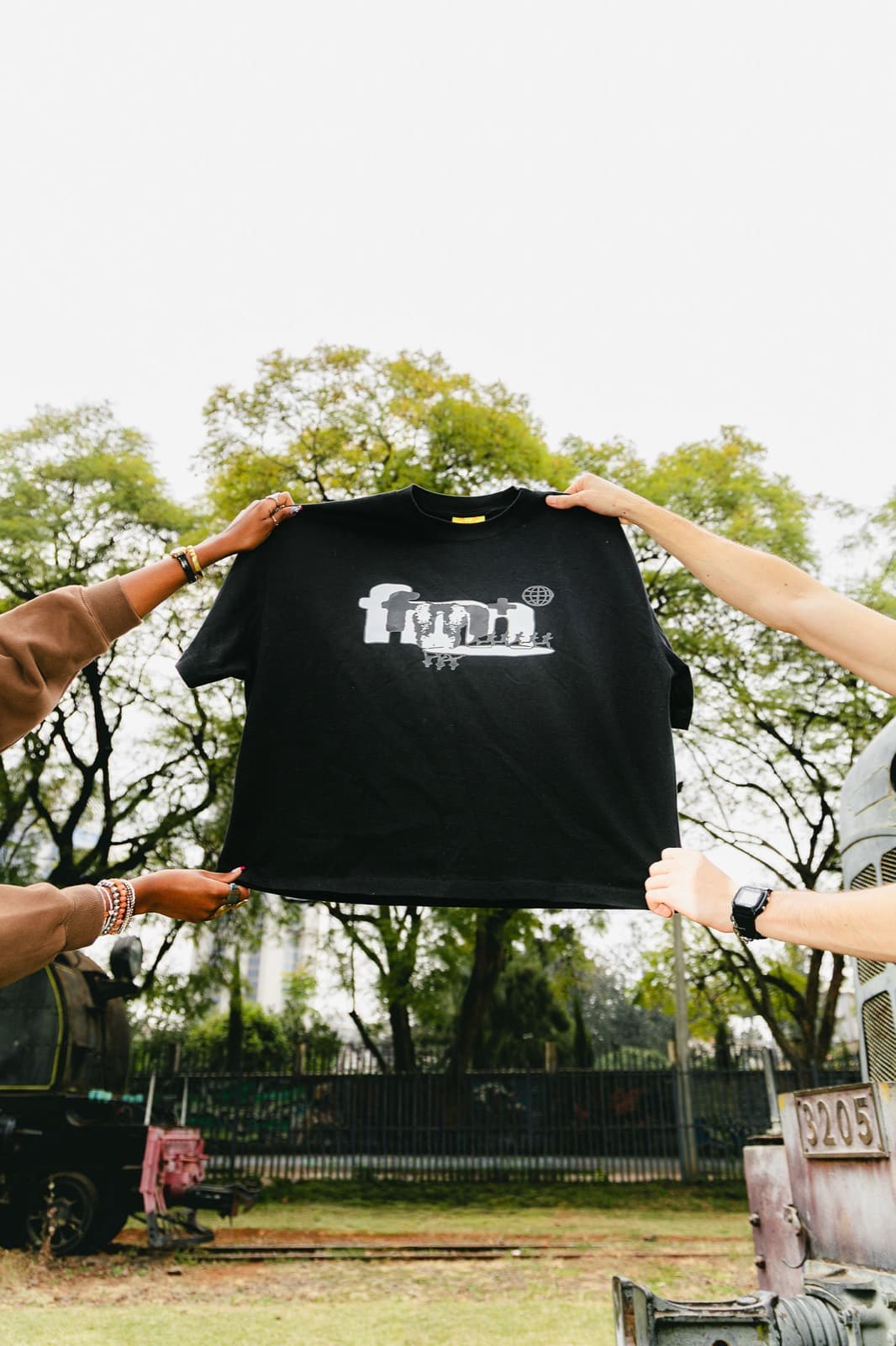 Four hands holding up the From NBO With Love Tee in Pitch Black / Mono against the trees.
